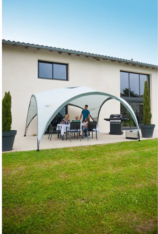 Een Coleman partytent met tafel en stoelen, naast een huis met een barbecue.