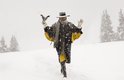 Man in lange zwarte jas met gele voering en sjaal, met hoed en twee pistolen in de sneeuw.