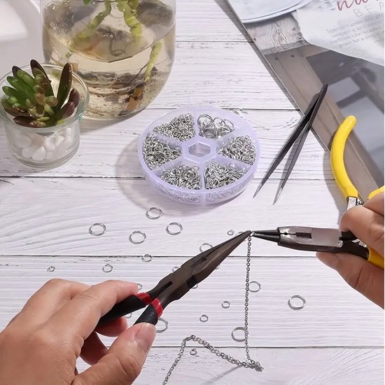 Handen die een zilverkleurige ketting maken met tangen en kleine zilverkleurige ringetjes, op een witte tafel met een opbergdoosje en plantje.