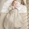 Baby in zandkleurige, gevoerde slaapzak met rits. Ligt op een laken met takjesprint in een ledikant.
