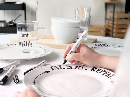 Une personne décore une assiette en porcelaine blanche avec l'inscription "EAT. SLEEP. REPEAT.". Un verre avec "BUT first MILK" est à côté.