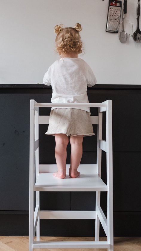 Kind met blonde knotjes in wit shirt en beige broek staat op witte leertoren, kijkt weg.