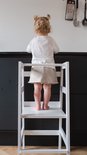 Kind met blonde knotjes in wit shirt en beige broek staat op witte leertoren, kijkt weg.