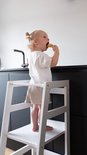 Kind met blonde knotjes in wit shirt en beige broek eet koekje op witte leertoren.