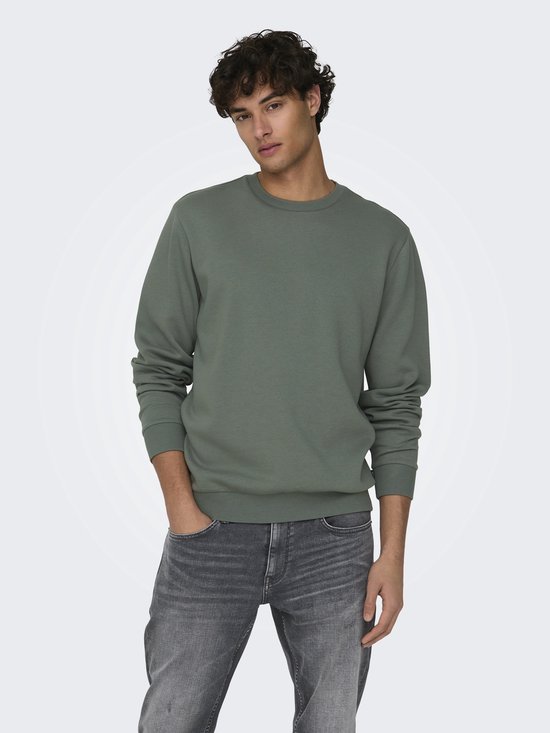 Een man met krullend haar draagt een mosgroene sweater met ronde hals en grijze jeans.