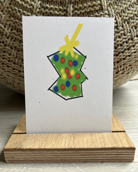 Een witte kaart met een groene kerstboom met rode en blauwe ballen en een gele strik, in een houten display.
