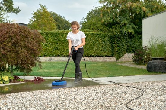 Vrouw met witte t-shirt en zwarte broek gebruikt een blauwe terrasreiniger met hogedrukreiniger in een tuin.