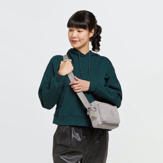 Vrouw met een donkergroene hoodie en zwarte leren broek, draagt een lichtgrijze Kipling schoudertas.
