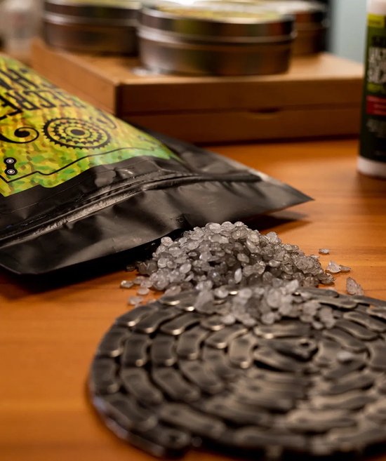 Zwarte zak met "SECRET CHAIN BLEND" etiket, grijze korrels en een fietsketting op een houten tafel.