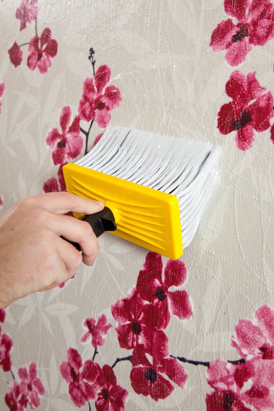 Hand met gele borstel brengt lijm aan op behang met bloemenprint.