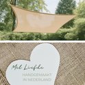 Beige schaduwdoek hangend tussen bomen. Een hartvormige kaart met 'Met Liefde Handgemaakt in Nederland' ligt op jute stof.