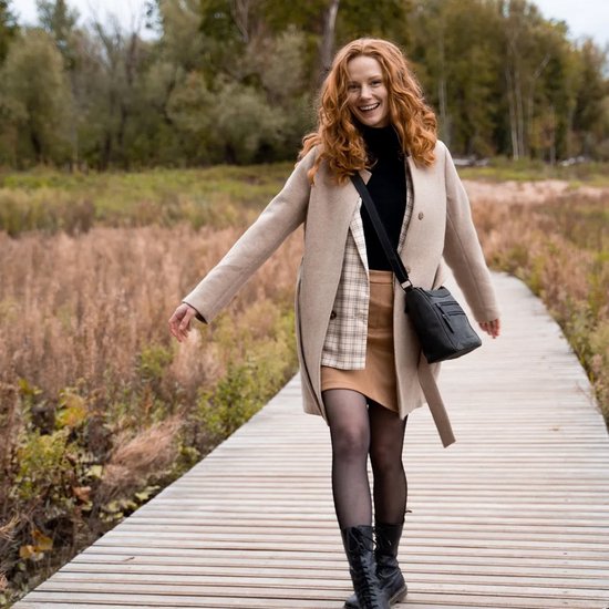 Roodharige vrouw met beige jas, zwarte coltrui, rok en laarzen, met zwarte schoudertas.