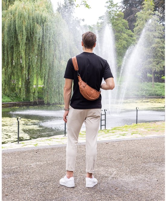 Man draagt een bruine leren crossbodytas op zijn rug, met een zwarte polo en beige broek, in een park met fonteinen.