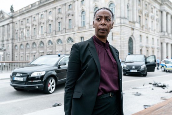 Een vrouw in een zwart pak en paarse blouse, met auto's en een gebouw op de achtergrond.