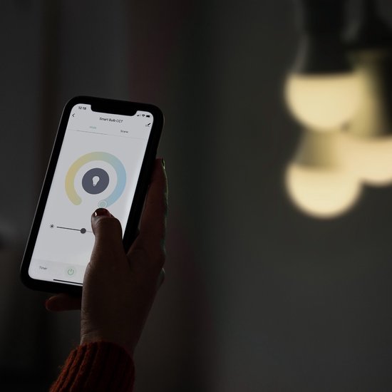 Hand houdt smartphone met de Hombli app, waarmee de 'Smart Bulb CCT' wordt bediend. Verlichting zacht op de achtergrond.