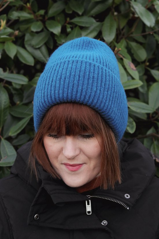 Vrouw met een blauwe gebreide muts en zwarte jas, met groene bladeren op de achtergrond.