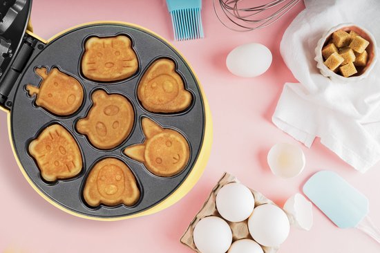 Gele wafelmaker van bovenaf gezien, gevuld met 7 ronde, bruine koekjes in diverse dierenvormpjes. Ingrediënten zoals eieren en suiker liggen ernaast.
