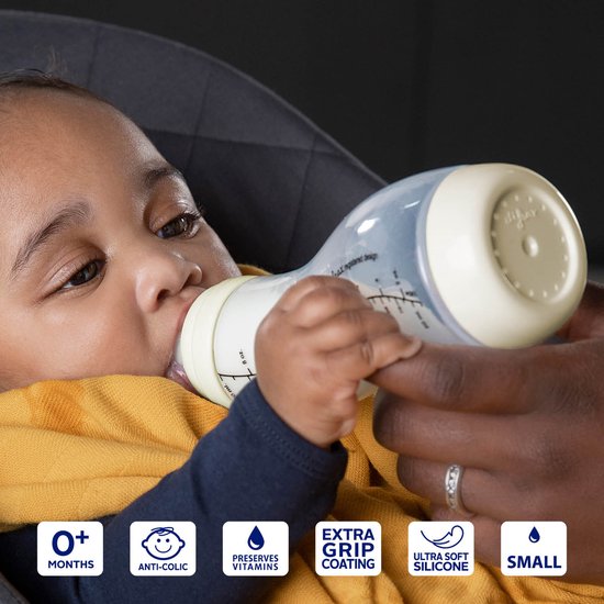 Bébé portant un pull jaune boit dans un biberon Difrax S couleur crème. Icônes: 0+ mois, anti-colique, vitamines, grip, silicone doux, petit.