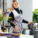 Vrouw met schort met paars-witte blokjes, met witte theedoek over haar schouder.