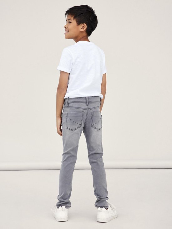 Jongen staat naar achteren gedraaid en draagt een grijze, aansluitende jeans en witte sneakers.