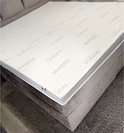 Witte matras met 'bamboo exclusive' opdruk op taupe bedbodem met grijs vloerkleed.