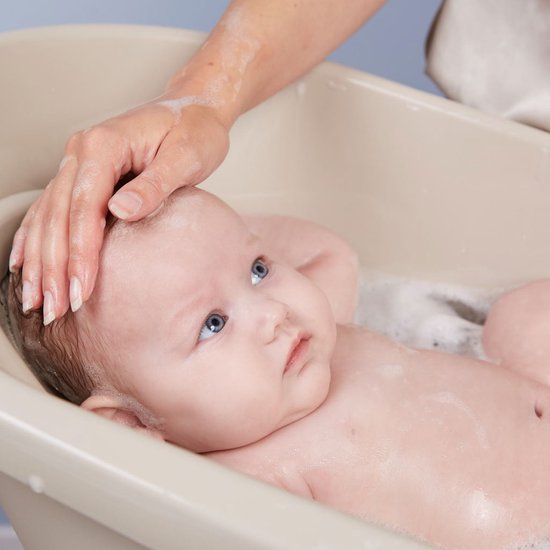 Een hand streelt het hoofd van een baby met blauwe ogen in een nat badje met schuim.