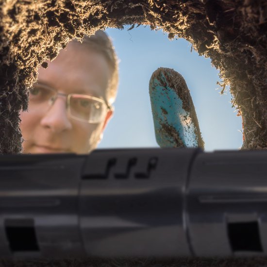 Perspectief vanuit een molshoop kijkend naar een persoon met bril en een schep, met de zwarte mollenklem op de voorgrond.