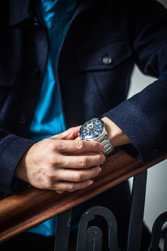 Close-up van een zilverkleurig herenhorloge met blauwe wijzerplaat en metalen band, gedragen door een model.