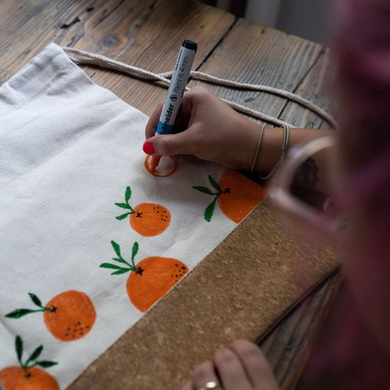Hand met oranje stift tekent sinaasappels op canvas tas.