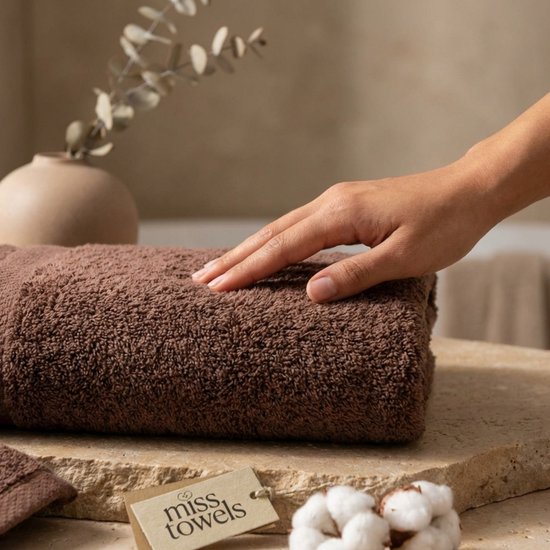 Hand rustend op een zachte, bruine handdoek met een 'miss towels' label en katoenbloemen op een stenen ondergrond.