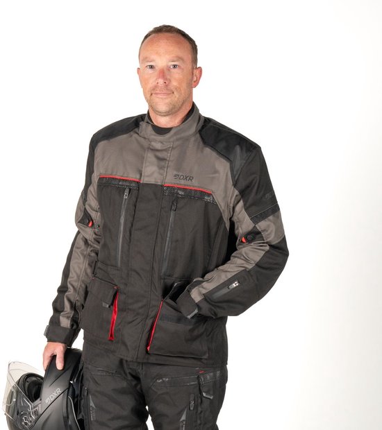 Man draagt een zwarte motorjas met donkergrijs en rode accenten en een motorbroek. Houdt een helm vast.