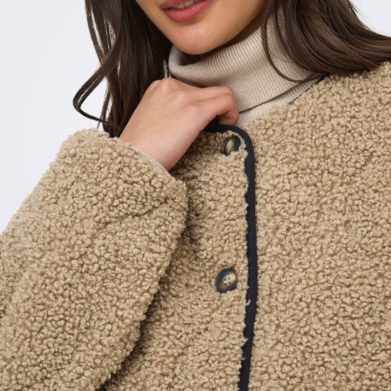 Close-up van een beige teddy jas met zwarte bies en knopen, gedragen over een coltrui.