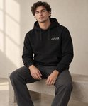 Man zit op een betonnen bankje, gekleed in een zwarte hoodie met COVAR-logo en grijze broek.