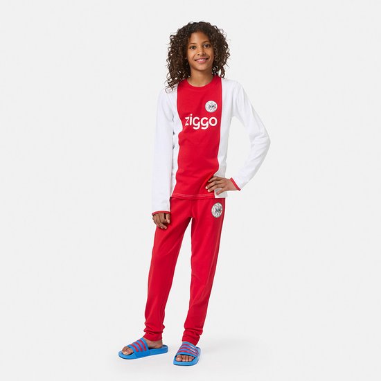Een jong meisje met krullend haar, draagt een Ajax pyjamaset met witte mouwen en een rood voorpand met 'ziggo' logo. De broek is rood.