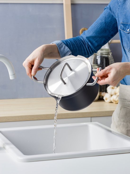 Een persoon met een blauwe spijkerblouse spoelt een matzwarte pan met RVS deksel af onder de kraan.
