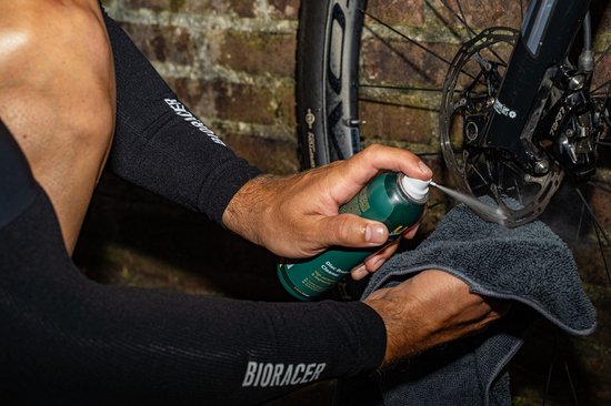Een hand spuit schijfreiniger op een fietsremschijf terwijl een andere hand een doek vasthoudt.