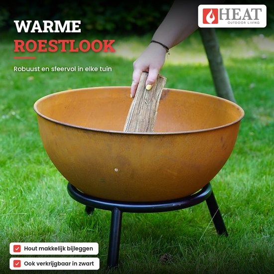 Roestkleurige vuurschaal op zwart onderstel in een tuin, hand plaatst houtblok erin.