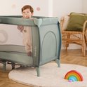Kind met fopspeen staat in een groen Lionelo reisbedje, op een houten vloer staat een regenboogspeeltje.