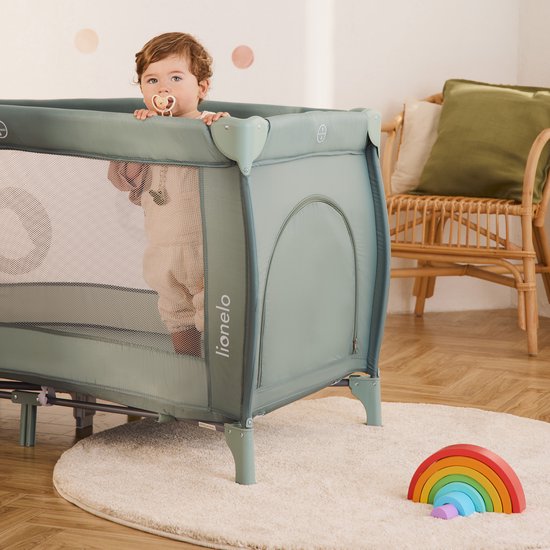 Kind met fopspeen staat in een groen Lionelo reisbedje, op een houten vloer staat een regenboogspeeltje.