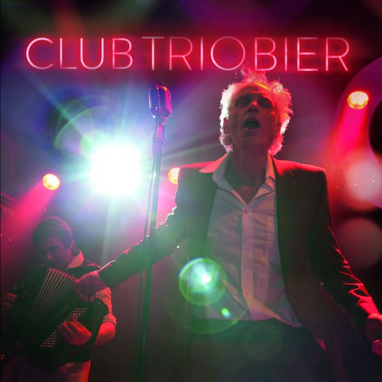 Een man met wapperend haar, zingend in een microfoon, met een accordeonist op de achtergrond. Neonlichten met de tekst CLUB TRIOBIER.