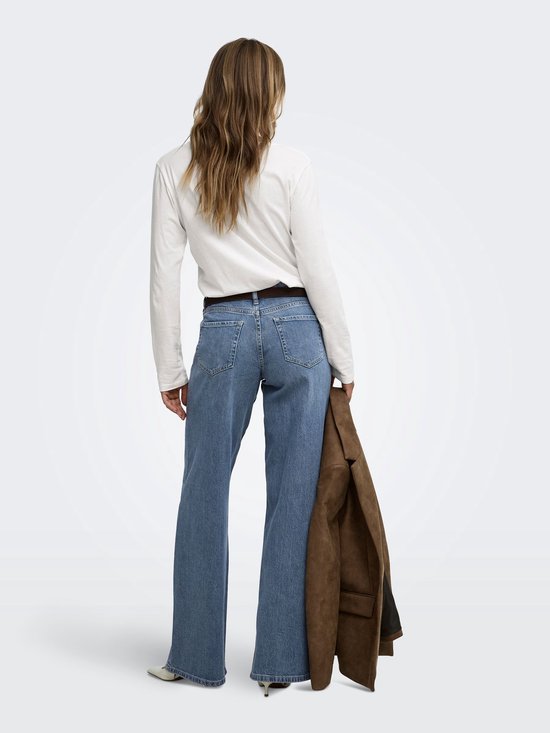 Model draagt een witte longsleeve, een spijkerbroek met wijde pijpen en een bruine blazer over de schouder.