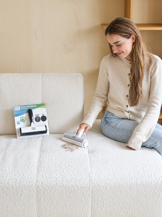 Femme portant un gilet beige et un jean utilise un rouleau anti-peluche sur un canapé blanc.