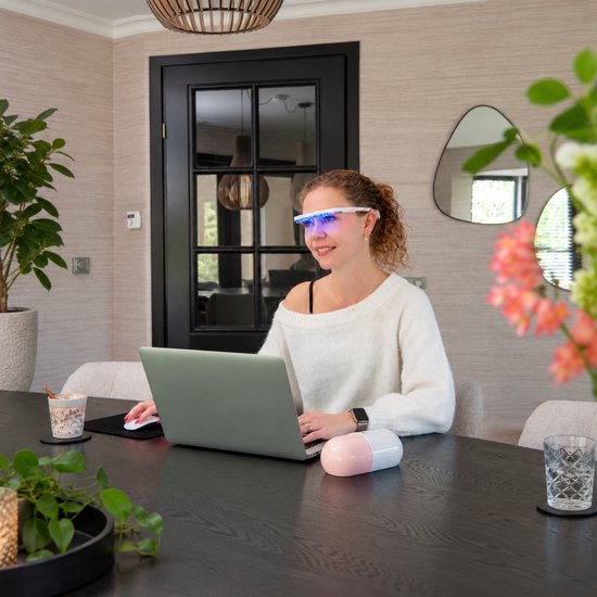 Vrouw met krullend haar draagt een witte AYO lichttherapiebril met blauw licht tijdens het werken op een laptop.