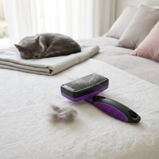 Paars-zwarte slickerborstel met grijze kattenharen op een wit laken, met een slapende grijze kat op de achtergrond.