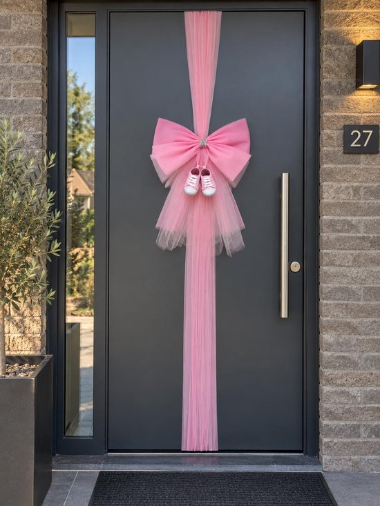 Grote roze strik van tule met glinsterende steentjes en babyschoentjes, versiert een donkergrijze voordeur.