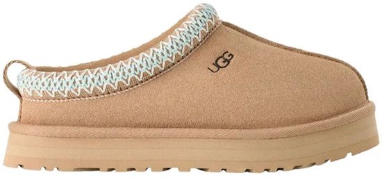 Bruine suède UGG-instapper met een dikke zool, blauw-wit vlechtwerk en het UGG-logo op de zijkant.