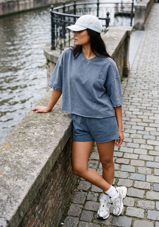 Model draagt een blauwe casual top met korte mouwen en een pet, staand bij een muur.