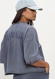 Model draagt een blauwe, geknipte top met korte mouwen en een pet, van achteren gezien.