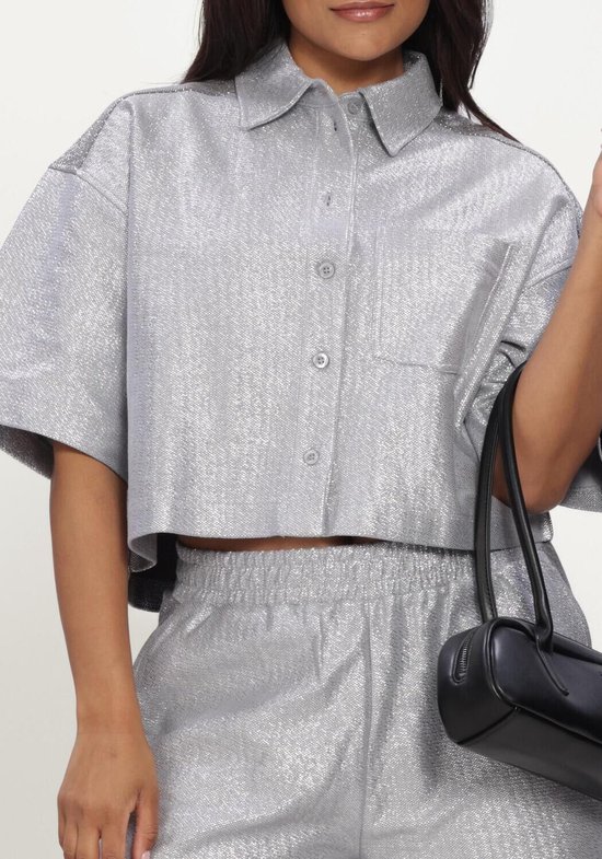 Buste van model in zilverkleurige, glanzende cropped blouse met korte mouwen en bijpassende shorts, met handtas.