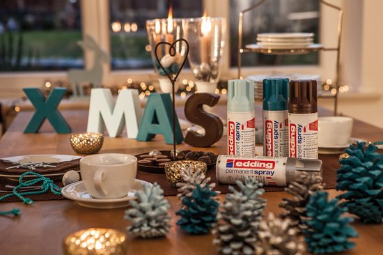 Kerstdecoratie met 'XMAS' letters, drie edding spuitbussen, kaarsen, dennenappels en tafelservies.